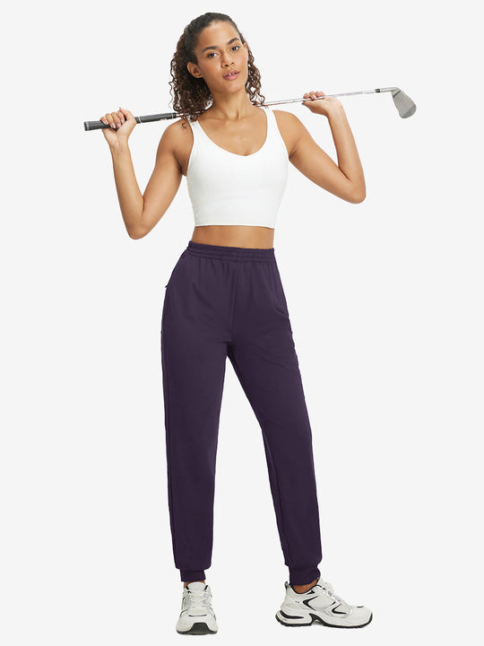 Model holding golf club behind neck while wearing lightweight women's stretchy joggers sweatpants in dark purple.